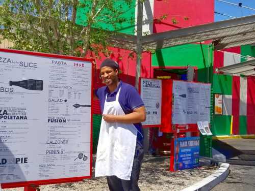 A smiling man in an apron stands in front of a colorful menu board outside a pizza restaurant.